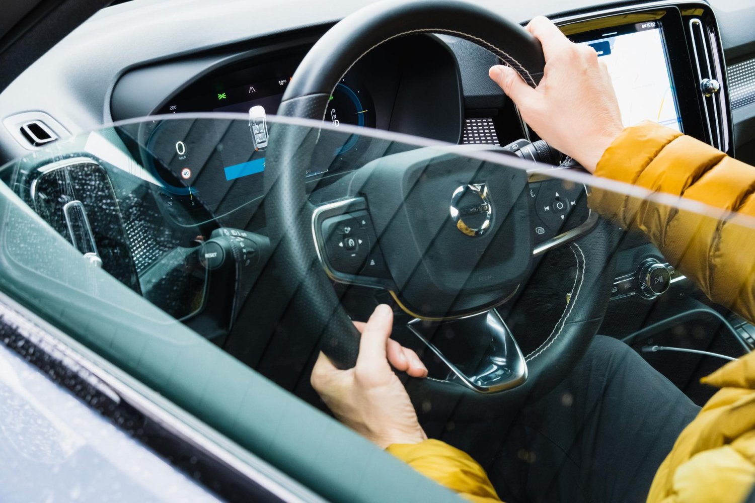 Nærbilde av rattet og dashbordet i en bil, sett utenfra. Man ser to hender som holder rattet.
