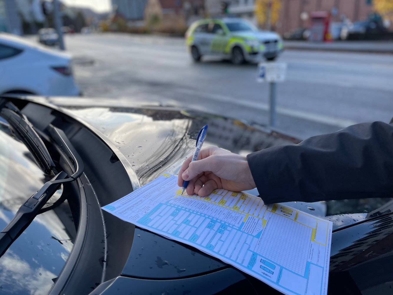 En mann fyller ut skadeskjema på et bilpanser. En politibil står parkert i bakgrunnen.