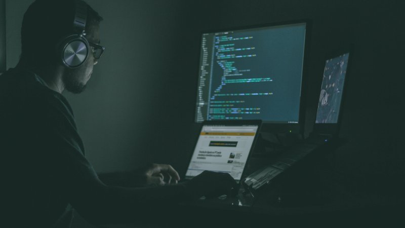 En mann sitter foran flere dataskjermer i et mørkt rom.