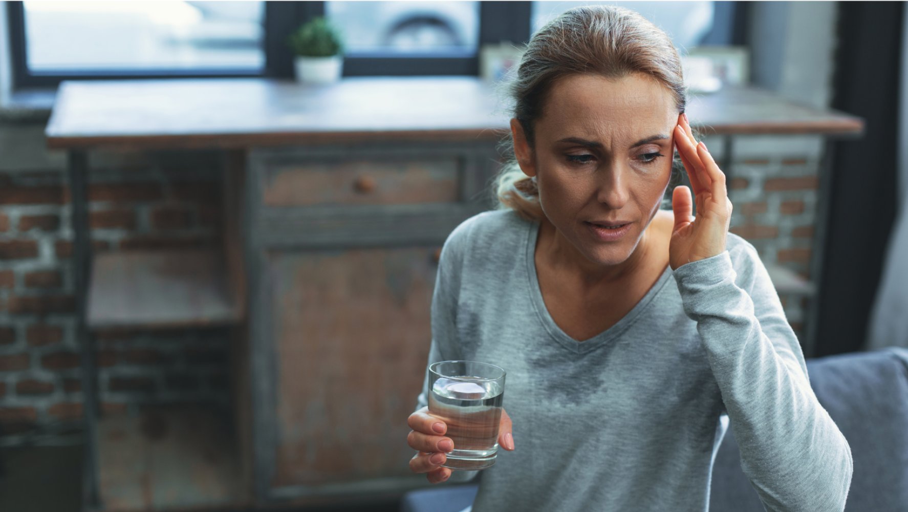En dame sitter med et glass vann i hånden mens hun tar seg til hodet.
