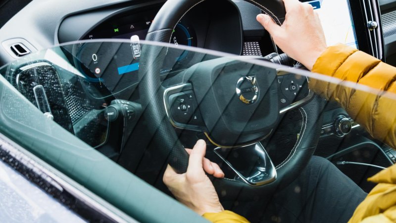 Nærbilde av rattet og dashbordet i en bil, sett utenfra. Man ser to hender som holder rattet.