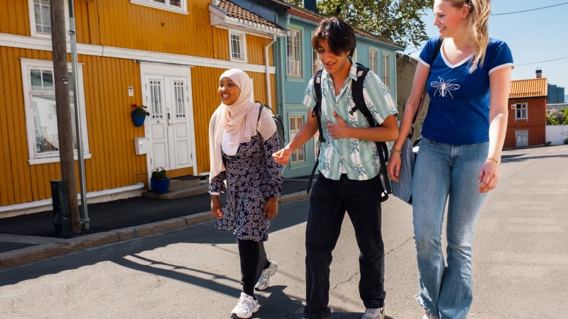 Tre unge mennesker, to kvinner og en mann, snakker og ler mens de går nedover gata. 