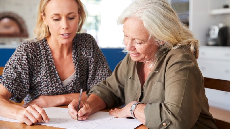 Bildet viser en eldre kvinne som signerer et papir, med hjelp fra en yngre kvinne.