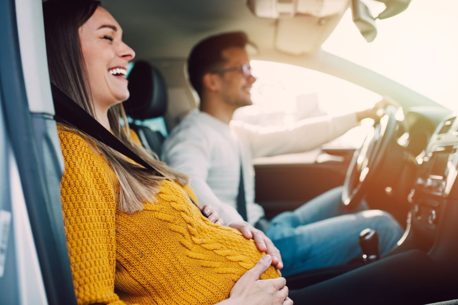 Bildet viser en gravid kvinne som sitter i bilen sammen med en mann. Hun smiler og holder seg på magen. 