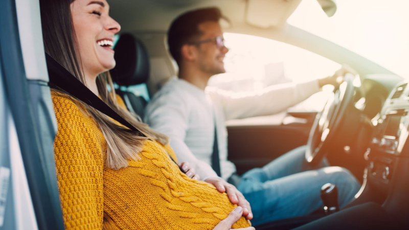 Bildet viser en gravid kvinne som sitter i bilen sammen med en mann. Hun smiler og holder seg på magen. 