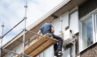 Bildet viser en håndverker som sitter i et stillas å jobber