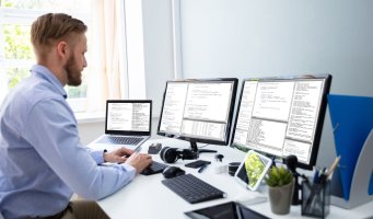 Bildet viser en mann som sitter foran tre skjermer på et kontor