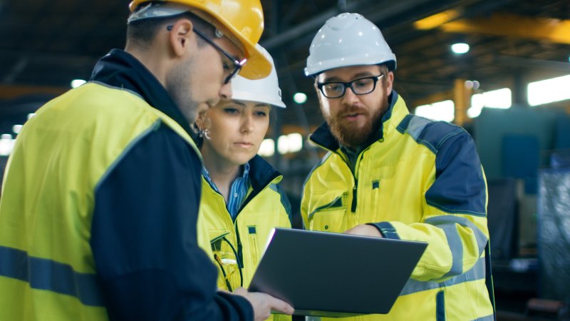Bildet viser tre arbeidere med refleksvest og hjelm på en byggeplass, som diskuterer noe som står på en skjerm.